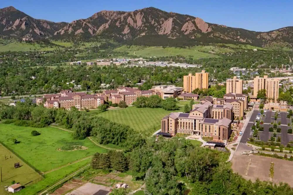 Aerial view of a lush green landscape in Boulder, featuring buildings and sprawling lawns, with a backdrop of rugged mountains, illustrating the natural environment where Boulder tree services and tree removal experts operate.